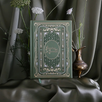 The Quirky Cup Collective reading journal in new sage colour way featured intricate gold and deep green foiling in a victorian-style aesthetic. The romance book inspired reading journal sits in front of a green drapey fabric background in front of a some white flowers and a brass vase.