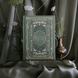 The Quirky Cup Collective reading journal in new sage colour way featured intricate gold and deep green foiling in a victorian-style aesthetic. The romance book inspired reading journal sits in front of a green drapey fabric background in front of a some white flowers and a brass vase.