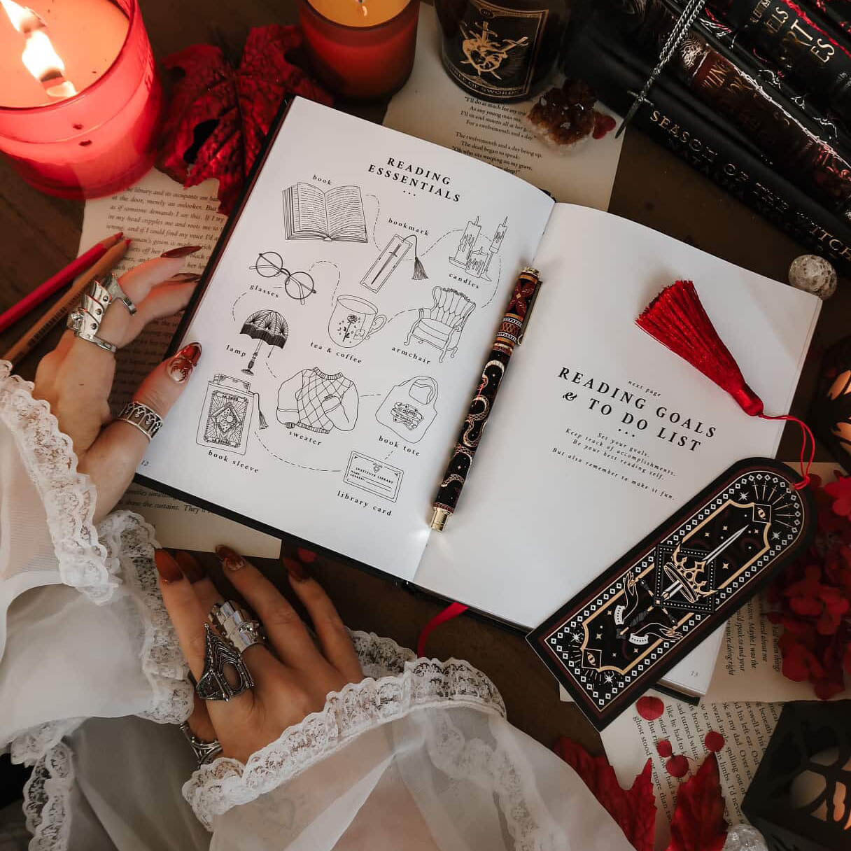A promo image for the Serpents & Swords Reading Journal by The Quirky Cup Collective. Open to a page section showing reading essentials, the journal is surrounded by a matching  black and red bookmark and snake embellished pen. Two hands with intricate rings wearing long white sleeves hold the book in a scene surrounded by candles and  foiled books.