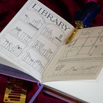 The gold Metal reading journal stencil ruler by The Quirky Cup Collective sits under an open reading journal showcasing the Library spread and is engraved with journaling symbols such as ratings, genre and a bookshelf on a deep pink red fabric background.