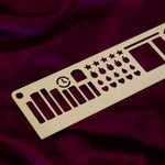 The gold Metal reading journal stencil ruler by The Quirky Cup Collective is engraved with journaling symbols such as ratings, genre and a bookshelf on a deep pink red fabric background.