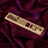 The gold Metal reading journal stencil ruler by The Quirky Cup Collective is engraved with journaling symbols such as ratings, genre and a bookshelf on a deep pink red fabric background.