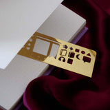 The gold Metal reading journal stencil ruler by The Quirky Cup Collective sits on an open reading journal and is engraved with journaling symbols such as ratings, genre and a bookshelf on a deep pink red fabric background.