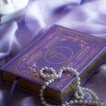 The cover of the highly decorative purple reading Journal by The Quirky Cup Collective sits surrounded by pearls in soft purple fabric. In a lilac colour, the romantasy inspired reading journal features a classic-inspired book ornamentation illustrations with gold and purple foil accents.