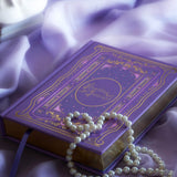 The cover of the highly decorative purple reading Journal by The Quirky Cup Collective sits surrounded by pearls in soft purple fabric. In a lilac colour, the romantasy inspired reading journal features a classic-inspired book ornamentation illustrations with gold and purple foil accents.