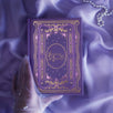 The cover of the highly decorative purple reading Journal by The Quirky Cup Collective sits surrounded by pearls in soft purple fabric, a hand in a sheer glove holding the book. In a soft lilac colour, the romantasy inspired reading journal features a classic-inspired book ornamentation illustrations with gold and purple foil accents.