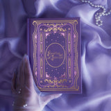 The cover of the highly decorative purple reading Journal by The Quirky Cup Collective sits surrounded by pearls in soft purple fabric, a hand in a sheer glove holding the book. In a soft lilac colour, the romantasy inspired reading journal features a classic-inspired book ornamentation illustrations with gold and purple foil accents.