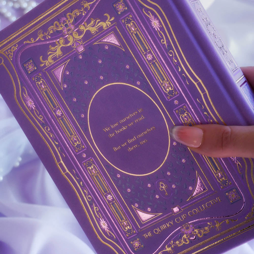 The back cover of the highly decorative purple reading Journal by The Quirky Cup Collective is held in front of a light purple background. In a lilac colour, the romantasy inspired reading journal features a classic-inspired book ornamentation illustrations with gold and purple foil accents.