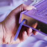 The edge of the highly decorative purple reading Journal by The Quirky Cup Collective is held by a hand. In a lilac colour, the romantasy inspired reading journal features a classic-inspired book ornamentation illustrations with gold and purple foil accents alongside a purple ribbon market and gold edge foiling.
