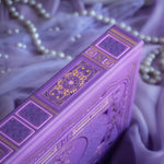 The spine of the highly decorative purple reading Journal by The Quirky Cup Collective sits surrounded by pearls in soft purple fabric. In a soft lilac colour, the spine featured classic-inspired ornamentation with gold foil accents for a romantasy inspired reading journal that feels bold and high end.