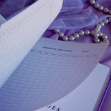 A spread of the Wisteria Lilac Reading journal by The Quirky Cup Collective sits on a soft lilac background with pearls and a dreamy, hazy aesthetic. The page shown is a Reading Tracker table.