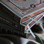 A detail image of the Sage trilogy of journals includes the standard journal, reading journal and reading companion by The Quirky Cup Collective in the sage Literati colour variation. Each journal features intricate gold foil detailing with gilded gold paper edges. The journals sit in front of a sage green fabric background.