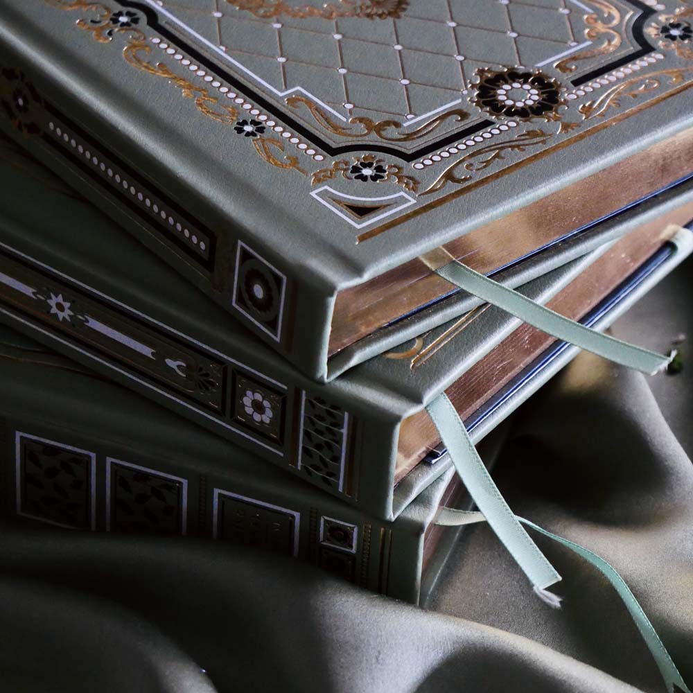 A detail image of the Sage trilogy of journals includes the standard journal, reading journal and reading companion by The Quirky Cup Collective in the sage Literati colour variation. Each journal features intricate gold foil detailing with gilded gold paper edges. The journals sit in front of a sage green fabric background.