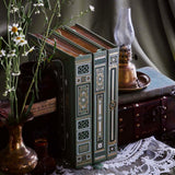 The Sage trilogy of journals includes the standard journal, reading journal and reading companion by The Quirky Cup Collective in the sage Literati colour variation. Each journal features intricate gold foil detailing and gold gilded page edges The journals sit together with spines out in front of a sage green fabric background alongside a candle stick and brass jug filled with flowers.