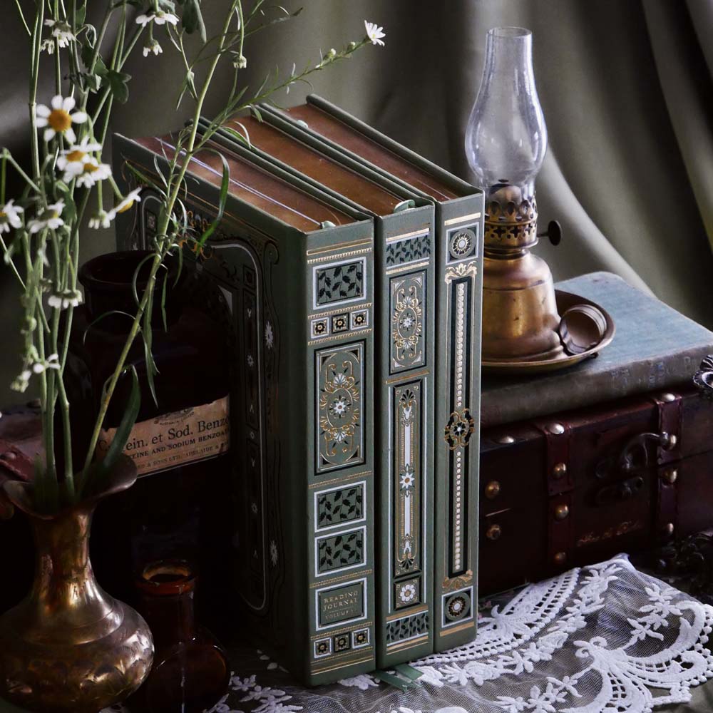 The Sage trilogy of journals includes the standard journal, reading journal and reading companion by The Quirky Cup Collective in the sage Literati colour variation. Each journal features intricate gold foil detailing and gold gilded page edges The journals sit together with spines out in front of a sage green fabric background alongside a candle stick and brass jug filled with flowers.