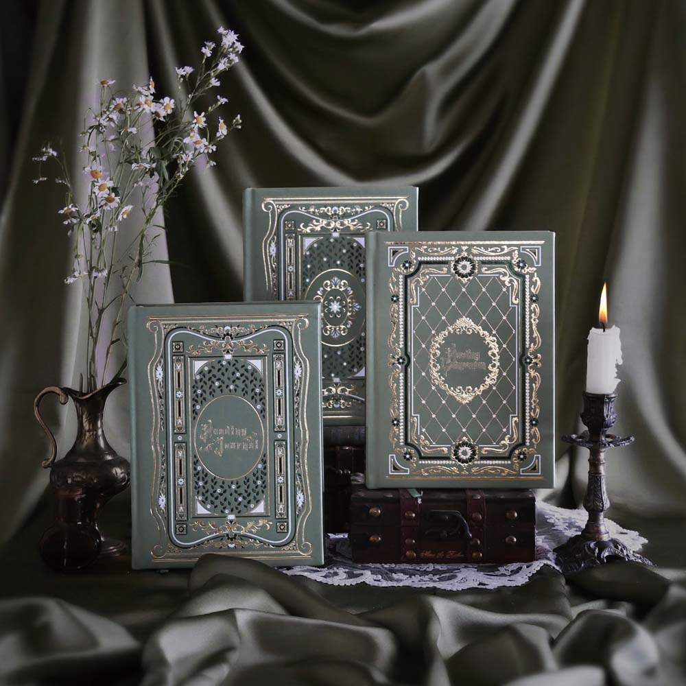 The Sage trilogy of journals includes the standard journal, reading journal and reading companion by The Quirky Cup Collective in the sage Literati colour variation. Each journal features intricate gold foil detailing with intricate patterns inspired b classical books. The journals sit in front of a sage green fabric background alongside a candle stick and brass jug filled with flowers.