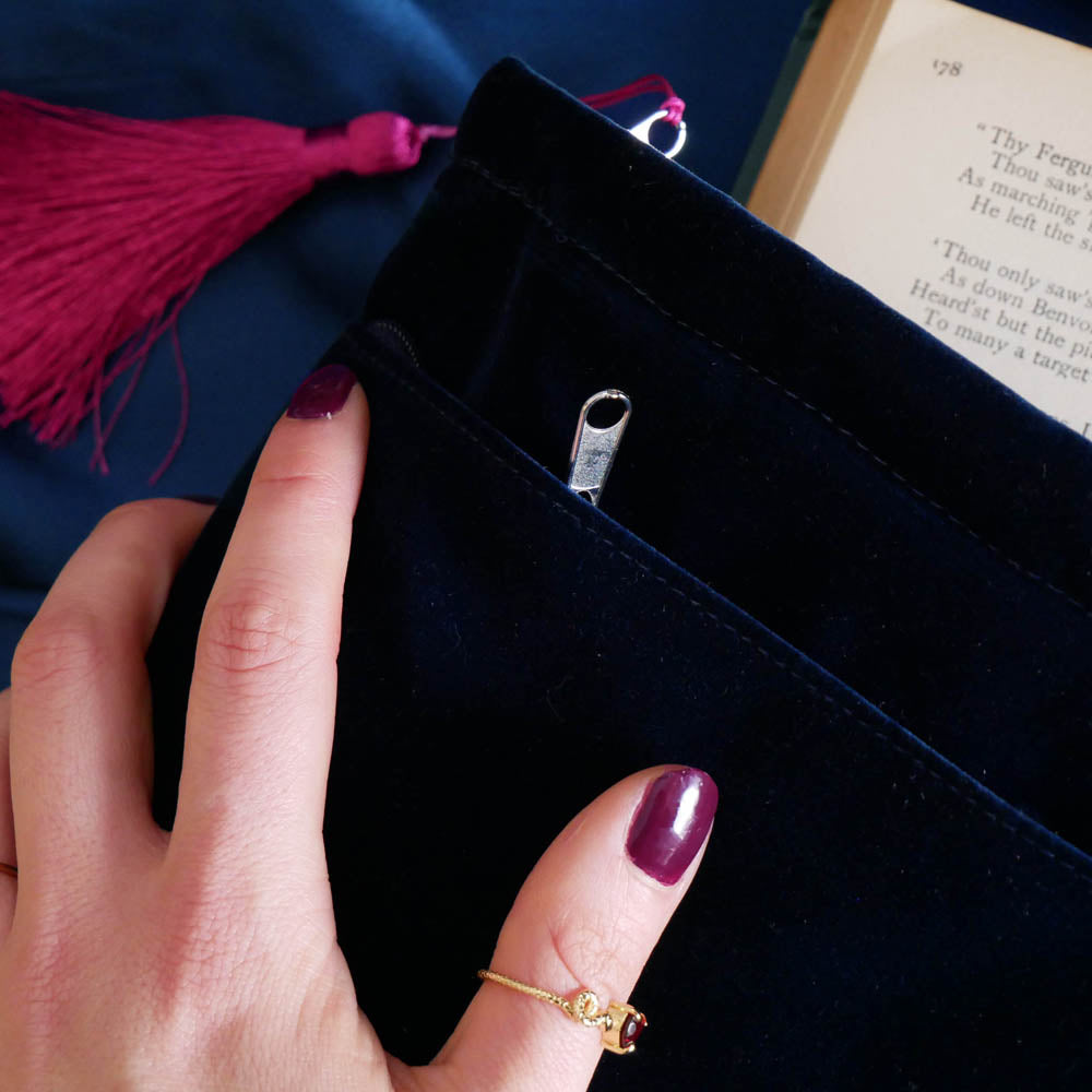 The back of the blue Serpents & Swords book sleeve. The velvet book case is a rich deep blue and features a bright pink tassel, the case held by a hand with purple nails over a blue fabric background.