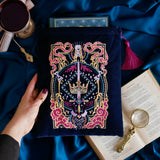 The blue Serpents & Swords book sleeve sits on a blue fabric background with a goblet, magnifying glass and classical book for a romantasy aesthetic. The book sleeve features intricate  embroidery of a snake, crown, sword and goblet motif in shades of pink, teal and gold. The book sleeve also has a bright pink tassle.