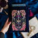 The blue Serpents & Swords book sleeve sits on a blue fabric background with a goblet, magnifying glass and classical book for a romantasy aesthetic. The book sleeve features intricate  embroidery of a snake, crown, sword and goblet motif in shades of pink, teal and gold. The book sleeve also has a bright pink tassle.