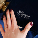 The back of the blue Serpents & Swords book sleeve sits on a blue fabric background. The dark blue velvet contrasts the crisp white logo of a key motif, a hand with purple nails holding the sleeve in front of a blue fabric background. 