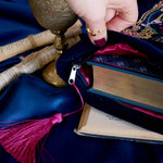 The blue Serpents & Swords book sleeve being held open with a large gilded book inside. The book sleeve features intricate  embroidery of a snake, crown, sword and goblet motif in shades of pink, teal and gold. The book sleeve also has a bright pink tassle and sits on a blue fabric background.