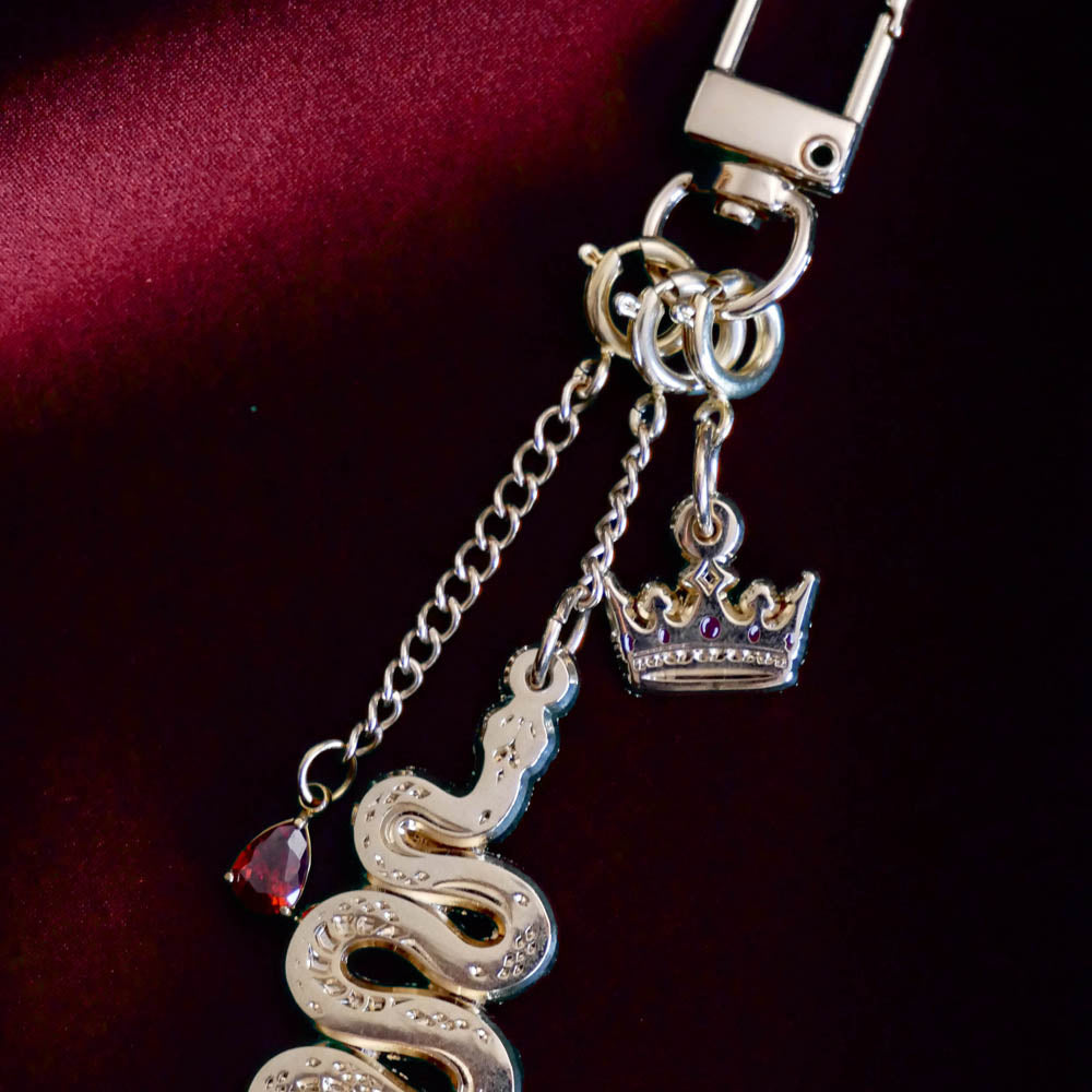 The detail of the gold Serpents & Swords charmlet by The Quirky Cup Collective sits on a red fabric background. The gold charm features three OTHER charms (that can be clipped/ interchanged)- one a small red gem, one a snake and one a crown with red detail. The charms can be mixed and matched for the perfect romantasy kindle aesthetic.