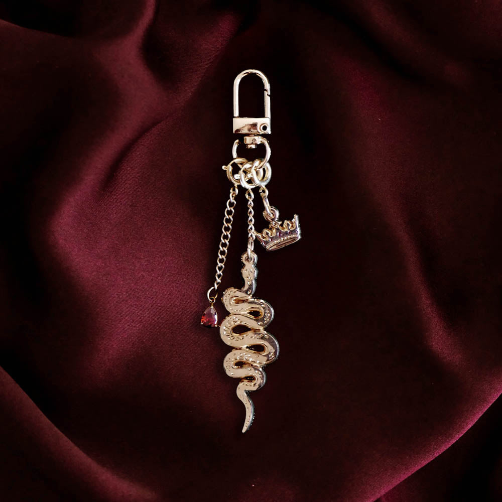 The gold Serpents & Swords charmlet by The Quirky Cup Collective sits on a red fabric background. The gold charm features three OTHER charms (that can be clipped/ interchanged)- one a small red gem, one a snake and one a crown with red detail. The charms can be mixed and matched for the perfect romantasy kindle aesthetic.