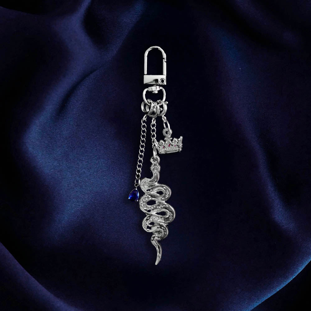 The Serpents & Swords charmlet by The Quirky Cup Collective sits on a deep blue fabric background. The silver charm features three OTHER charms (that can be clipped/ interchanged)- one a small blue gem, one a snake and one a crown with red detail. The charms can be mixed and matched for the perfect romantasy kindle aesthetic.
