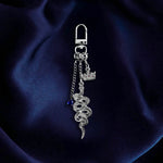 The Serpents & Swords charmlet by The Quirky Cup Collective sits on a deep blue fabric background. The silver charm features three OTHER charms (that can be clipped/ interchanged)- one a small blue gem, one a snake and one a crown with red detail. The charms can be mixed and matched for the perfect romantasy kindle aesthetic.