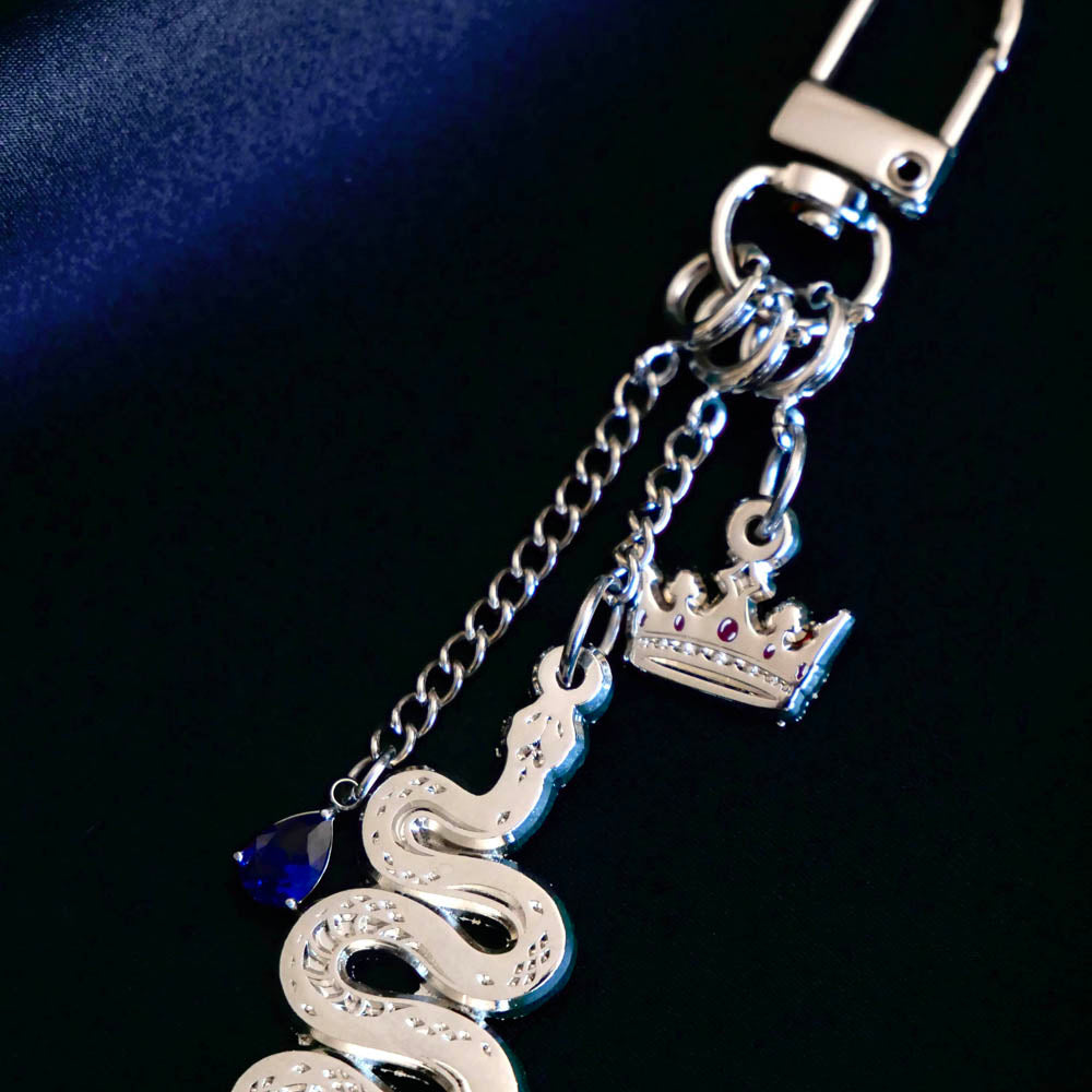 The detail of the silver Serpents & Swords charmlet by The Quirky Cup Collective sits on a deep blue fabric background. The silver charm features three OTHER charms (that can be clipped/ interchanged)- one a small blue gem, one a snake and one a crown with red detail. The charms can be mixed and matched for the perfect romantasy kindle aesthetic.