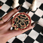 The black oath Breaker clingie (think a bookish enamel pop socket!) is held in front of a black and white chess board. The design centres on a crown, sword and snake motif with two hands at the bottom. The mostly circular (the bottom has a 'notch' Clingie is in shades of red black and white alongside the gold enamel.