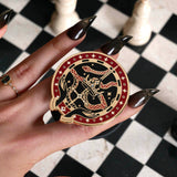 The black oath Breaker clingie (think a bookish enamel pop socket!) is held in front of a black and white chess board. The design centres on a crown, sword and snake motif with two hands at the bottom. The mostly circular (the bottom has a 'notch' Clingie is in shades of red black and white alongside the gold enamel.