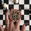 The black oath Breaker clingie (think a bookish enamel pop socket!) is held in front of a black and white chess board. The design centres on a crown, sword and snake motif with two hands at the bottom. The mostly circular (the bottom has a 'notch' Clingie is in shades of red black and white alongside the gold enamel.