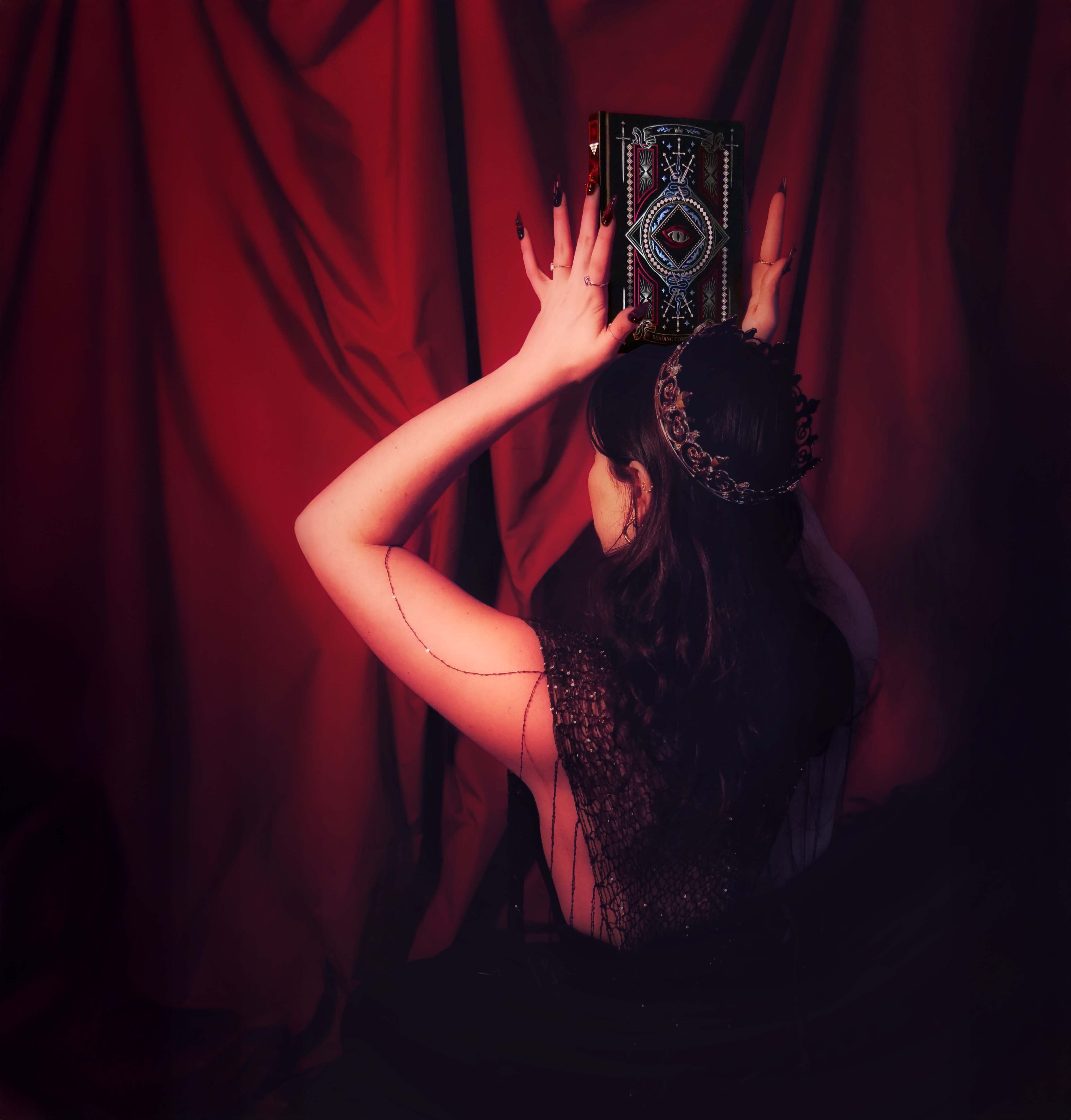 A woman wearing a crown holds an intricate journal by The Quirky Cup Collection above her head against a red curtain backdrop. The foiled reading journal has a dragon eye motif alongside swords and ornate patterns.