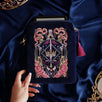 The navy blue Serpents & Swords eReader case features an intricate embroidery in shades of bright pink,, hold, teal and light pink. Motifs of snakes, crowns, swords, eyes and hands make for the perfect romantasy inspired gift. The velvet case with pink ribbon and wrist strap sits on a royal blue fabric background, a kindle poking out the top.