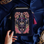The navy blue Serpents & Swords eReader case features an intricate embroidery in shades of bright pink,, hold, teal and light pink. Motifs of snakes, crowns, swords, eyes and hands make for the perfect romantasy inspired gift. The velvet case with pink ribbon and wrist strap sits on a royal blue fabric background, a kindle poking out the top.