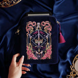 The navy blue Serpents & Swords eReader case features an intricate embroidery in shades of bright pink,, hold, teal and light pink. Motifs of snakes, crowns, swords, eyes and hands make for the perfect romantasy inspired gift. The velvet case with pink ribbon and wrist strap sits on a royal blue fabric background, a kindle poking out the top.