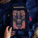 The navy blue Serpents & Swords eReader case features an intricate embroidery in shades of bright pink,, hold, teal and light pink. Motifs of snakes, crowns, swords, eyes and hands make for the perfect romantasy inspired gift. The velvet case with pink ribbon and wrist strap sits on a royal blue fabric background, a kindle poking out the top.