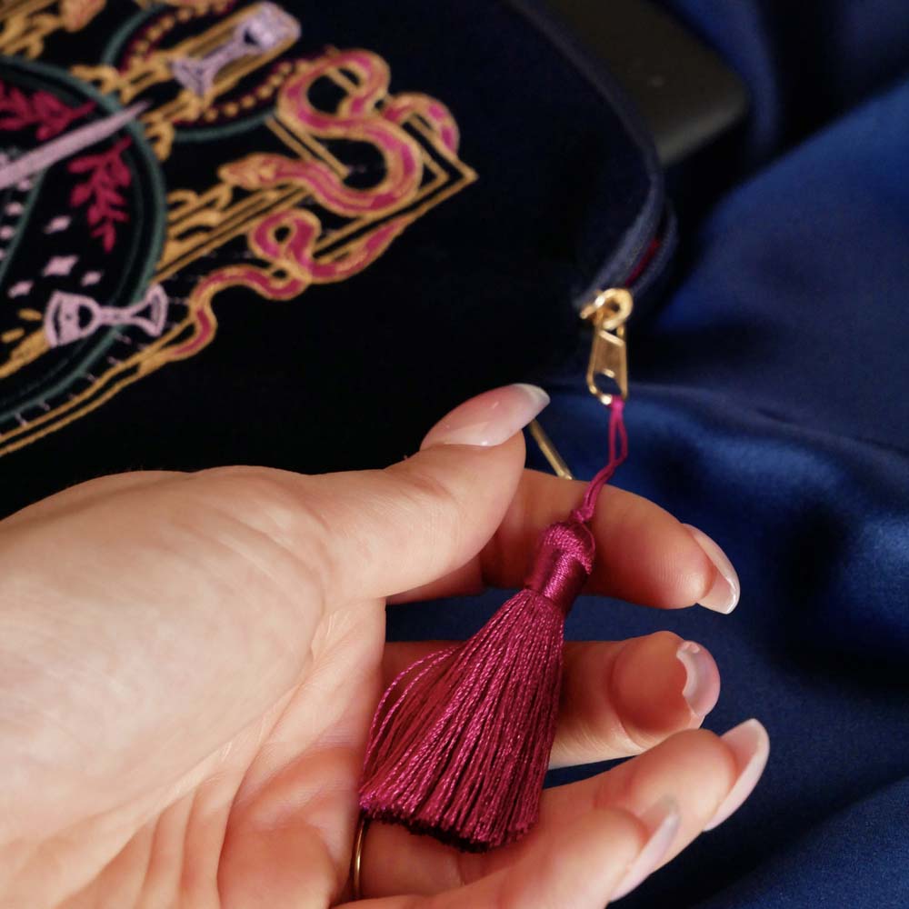 A detail of the  navy blue Serpents & Swords eReader case features an intricate embroidery in shades of bright pink,, hold, teal and light pink. Motifs of snakes, crowns, swords, eyes and hands make for the perfect romantasy inspired gift. The velvet case with pink tassel and wrist strap sits on a royal blue fabric background.