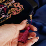 A detail of the  navy blue Serpents & Swords eReader case features an intricate embroidery in shades of bright pink,, hold, teal and light pink. Motifs of snakes, crowns, swords, eyes and hands make for the perfect romantasy inspired gift. The velvet case with pink tassel and wrist strap sits on a royal blue fabric background.