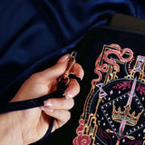 A detail of the  navy blue Serpents & Swords eReader case features an intricate embroidery in shades of bright pink,, hold, teal and light pink. Motifs of snakes, crowns, swords, eyes and hands make for the perfect romantasy inspired gift. The velvet case with pink tassel and wrist strap sits on a royal blue fabric background, a hand around the strap.