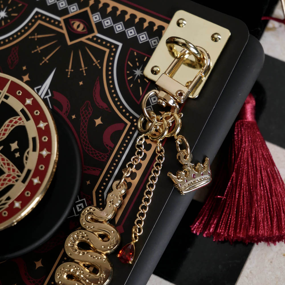 A detail of the gold charm anchor attached to a Kindle with the serpents & Swords charmlet and matching Clingie and eReader insert sticker.  The rectangular anchor sticks the the charmlet (which features a gold snake, crown and small red gem dangle) to the device. The  clingie and eReader insert sticker design centres on a crown, sword and snake motifs in red, gold and black tones.