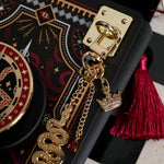 A detail of the gold charm anchor attached to a Kindle with the serpents & Swords charmlet and matching Clingie and eReader insert sticker.  The rectangular anchor sticks the the charmlet (which features a gold snake, crown and small red gem dangle) to the device. The  clingie and eReader insert sticker design centres on a crown, sword and snake motifs in red, gold and black tones.