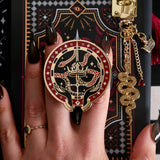 The black oath Breaker clingie is held attached to a kindle with matching charmlet and eReader insert sticker. The clingie (think a fantasy inspired enamel pop socket!) design centres on a crown, sword and snake motif with two hands at the bottom. The mostly circular (the bottom has a 'notch' Clingie is in shades of red, black and white alongside the gold enamel. The charmlet shows a gold snake, crown and small red gem dangle.