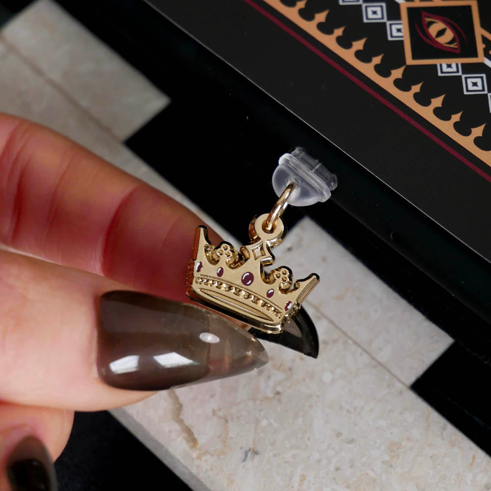 The gold Gilded Heir dust plug is essential romantasy aesthetic kindle decor- it is shown here in a eReader over a chessboard. The dust plug features a small gold crown motif with red details.
