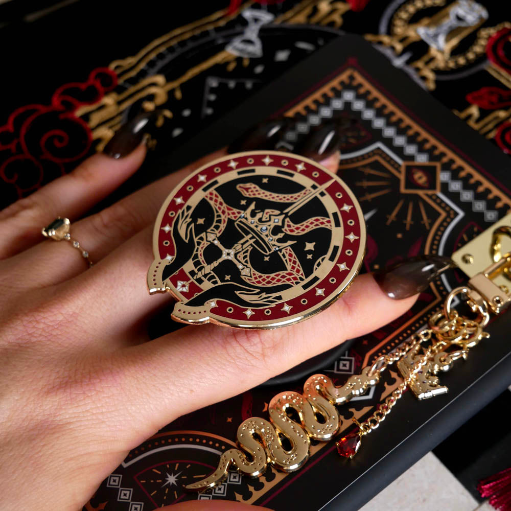 A detail of the Serpents & Swords eReader decor bundle. On a chess board on a black background a hand holds the oath Breaker clingie (think an enamel pop socket!), attached to a kindle with matching eReader sticker insert. It sits on the matching eReader case and has a matching charmlet attached. The range features snake, crown and sword motifs and is black with gold, red and white detailing.