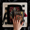 A detail of the Serpents & Swords eReader decor bundle. On a chess board on a black background a hand holds the oath Breaker clingie (think an enamel pop socket!), attached to a kindle with matching eReader sticker insert. It sits on the matching eReader case and has a matching charmlet attached. The range features snake, crown and sword motifs and is black with gold, red and white detailing.