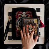 A detail of the Serpents & Swords eReader decor bundle. On a chess board on a black background a hand holds the oath Breaker clingie (think an enamel pop socket!), attached to a kindle with matching eReader sticker insert. It sits on the matching eReader case and has a matching charmlet attached. The range features snake, crown and sword motifs and is black with gold, red and white detailing.