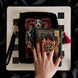 A detail of the Serpents & Swords eReader decor bundle. On a chess board on a black background a hand holds the oath Breaker clingie (think an enamel pop socket!), attached to a kindle with matching eReader sticker insert. It sits on the matching eReader case and has a matching charmlet attached. The range features snake, crown and sword motifs and is black with gold, red and white detailing.