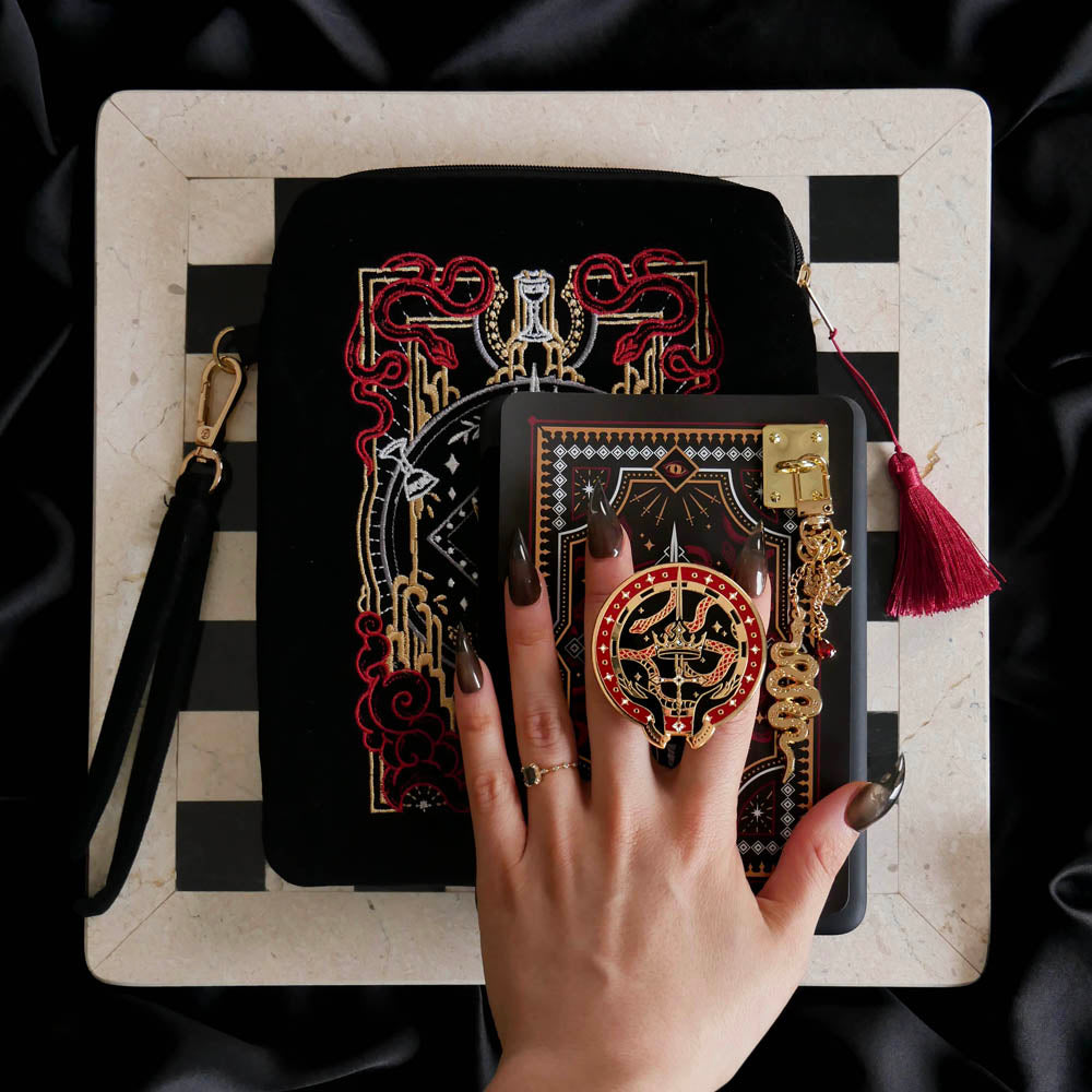 A detail of the Serpents & Swords eReader decor bundle. On a chess board on a black background a hand holds the oath Breaker clingie (think an enamel pop socket!), attached to a kindle with matching eReader sticker insert. It sits on the matching eReader case and has a matching charmlet attached. The range features snake, crown and sword motifs and is black with gold, red and white detailing.